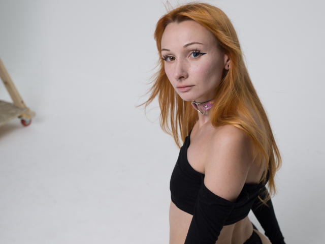 HannaCool with red hair, black off-shoulder top, pink collar, looking at camera, small tits, light skin, slight smile, standing, white background, soft lighting, slight wind in hair.