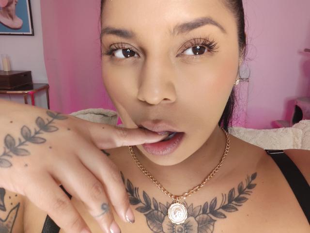 Chili, com tatuagens no peito e na mão, chupa o dedo, olhos meio abertos, colar de ouro, fundo rosa.