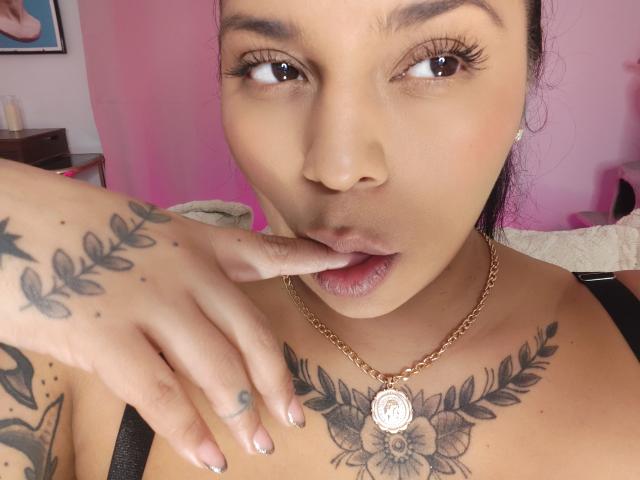 Chili, with big tits and tattoos on her chest and hand, seductively bites her finger while wearing a gold necklace.