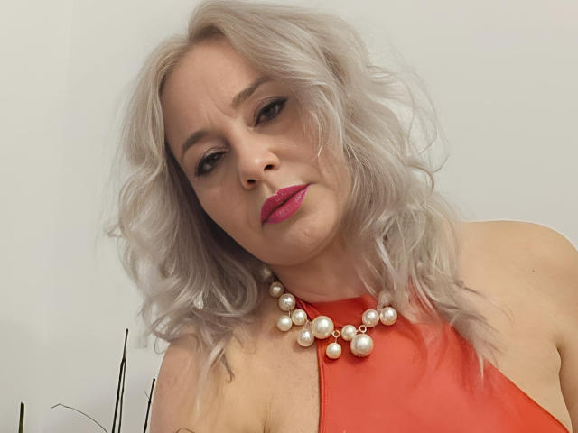 EmilyLowe69 with wavy white hair, pink lipstick, and red dress, wearing a pearl necklace, looking seductively at the camera, showing her tits in the photo.