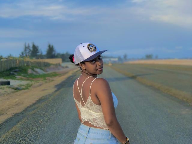 Laulah with dark skin, small tits, wearing a white lace bralette, high-waisted blue jeans, and a white baseball cap, standing on a gravel road, looking back.