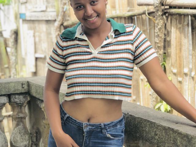 Laulah with light brown skin, small breasts, and a cute smile, wearing a striped crop top and blue jeans, leaning on a stone balcony, showing her flat stomach.