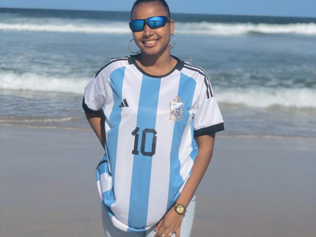 Laulah with dark skin, blue sunglasses, hoop earrings, and a blue-and-white Argentine soccer jersey number 10, standing on a beach, smiling, wearing a gold watch.