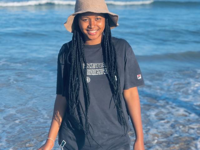 Laulah with dark skin, long black braids, smiling, wearing a beige bucket hat and black t-shirt, standing in shallow water. Her tits are small, water touching her legs.