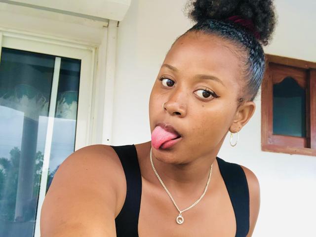 Laulah with dark brown skin, curly black hair in a bun, tongue out, wearing a black tank top, small hoop earrings, and a silver necklace.