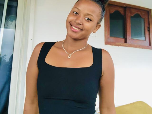 Laulah with dark skin and small tits, smiling, wearing a black tank top, silver necklace, and hoop earrings, standing indoors, her hair in a bun.