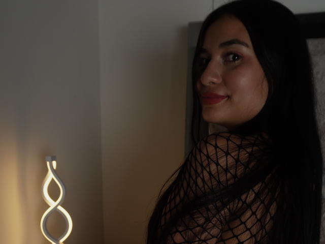 AliceNova met lang zwart haar, lachend, met een top van netstof, waarbij haar borsten zichtbaar zijn, kijkt over haar schouder, staat in de buurt van een gloeiende wandlamp, donkere kamer, rode lippenstift..