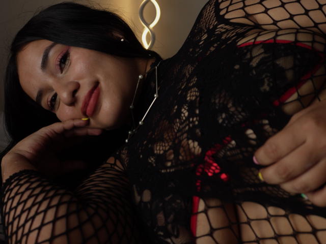 AliceNova in fishnet top, showing her tits, with red bra visible, black hair, pink eyeshadow, yellow nail, touching face, smiling, ringed fingers, necklace, dim light.