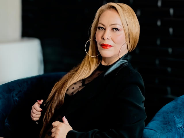 TheSweetHoney with blonde hair, red lipstick, black sheer top, polka dot pattern, large hoop earrings, sitting on blue velvet chair, black background, holding black jacket.