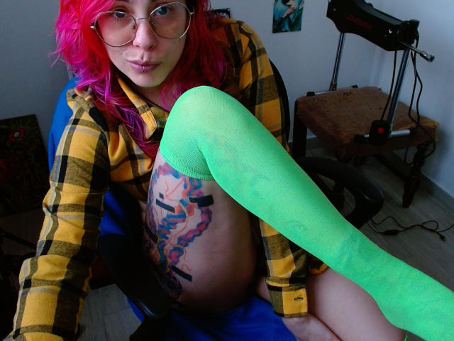 NatashaLau, con cabello rosa, gafas, una camisa amarilla a cuadros y medias altas de color verde neón, mostrando un tatuaje colorido en su lado izquierdo, sentada en una silla azul..