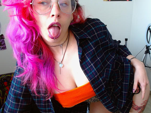 NatashaLau, com cabelo rosa, língua de fora, usando óculos, camisa xadrez, blusa laranja e colar, mostrando os seios. Ela está inclinada para frente, com a mão na coxa, e parece estar se divertindo..