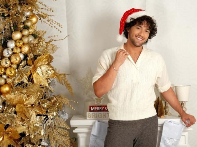 FreyrStyle69 with curly brown hair, smiling, wearing a red Santa hat, white knit shirt, and gray pants, standing next to a gold and silver Christmas tree.