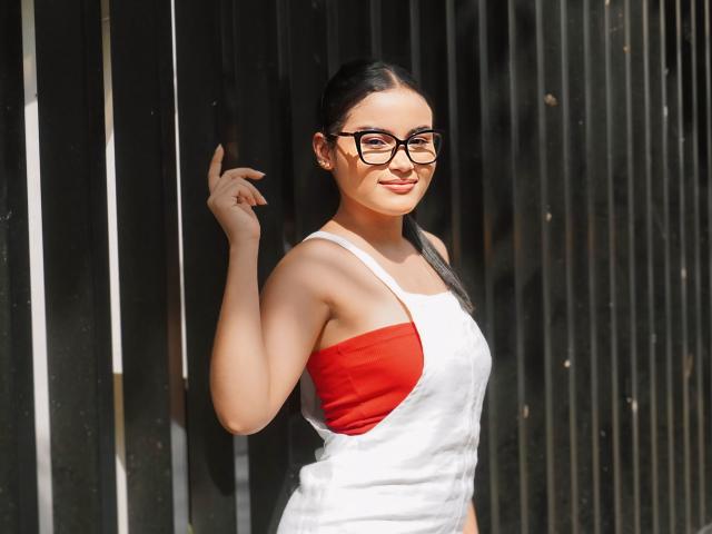 MiiaPalmer with glasses, dark hair in a ponytail, white tank top with red side, showing her tits, standing against a black vertical metal wall, smiling slightly.