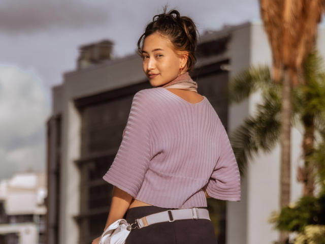 NofaceNice with brown hair in a messy bun, wearing a purple ribbed sweater and black pants, looking over her shoulder with a slight smile, standing outdoors.