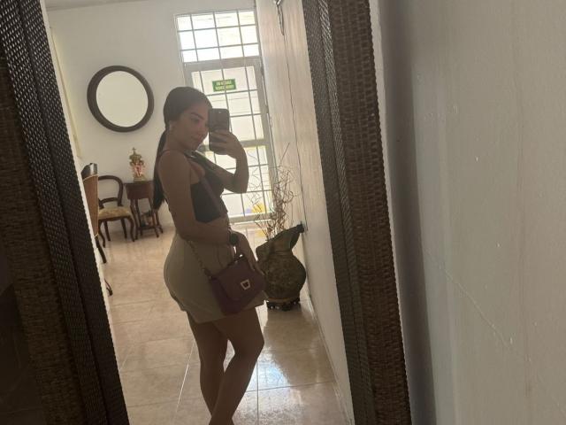 SharonKlart, reflecting in a mirror, wearing a black top and white booty shorts, holding a brown purse, with her ass and boobs slightly visible, long dark hair.