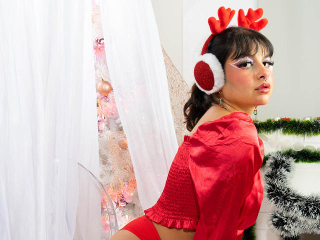 LaraAhB com chifres de rena vermelhos, uma cropped vermelha e calcinha vermelha, mostrando o bumbum. Ela está olhando para trás, com cortinas brancas e uma árvore de Natal decorada ao fundo..