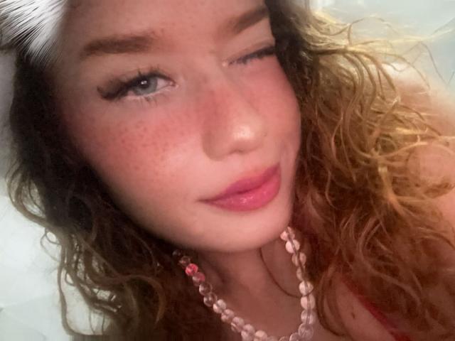 AkiraZanoli with wavy blonde hair, one eye closed, pink lips, freckles, and a pink beaded necklace, looking at the camera with a slight smile. Her tits are not visible.