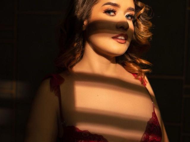 AkiraZanoli with curly brown hair, wearing red lace bra, has sunlight casting shadow across her tits and face, looking up with a slight smile, in warm light.