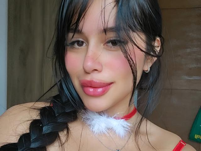 SophyLeon has black hair in a braid, pink lips, and a red choker with white fur. She's smiling with a slight blush on her cheeks. Her tits are partially visible.