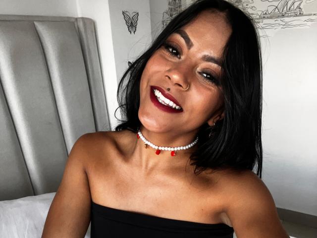 Amberv has dark skin, black hair, and is smiling with red lipstick. She's wearing a black strapless top and a red-beaded necklace. Her tits are small.