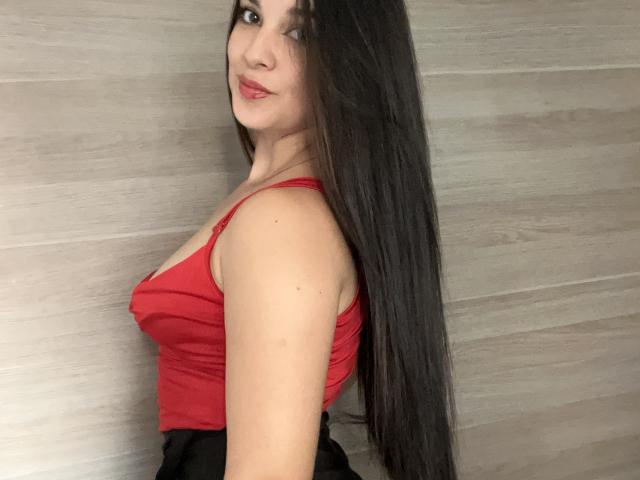 Katelyn Allen, met lang donker haar, een rode tanktop, waarbij haar borsten zichtbaar zijn, een zwarte rok, lachend, kijkend over haar schouder, lichte huid, een houten muur op de achtergrond, een lichte blos op haar wangen..
