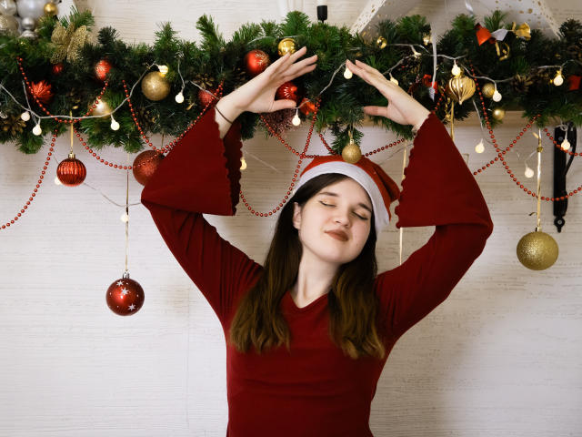 BonnieFlower con los ojos cerrados, vistiendo un vestido rojo de manga larga y un gorro de Papá Noel, con los brazos levantados, tocando una guirnalda navideña con adornos rojos y dorados, una pared blanca al fondo..