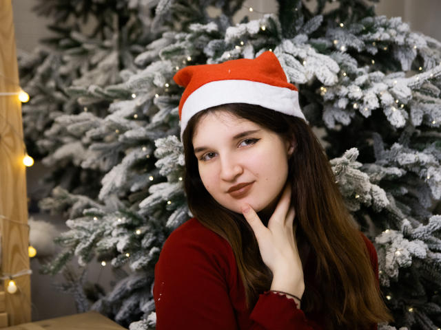 BonnieFlower, con cabello castaño largo, un gorro rojo de Papá Noel y un suéter rojo, tocándose la barbilla, de pie frente a un árbol de Navidad cubierto de nieve con luces pequeñas..