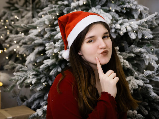 BonnieFlower with long brown hair, wearing a red Santa hat and red sweater, finger to lips, standing in front of a snow-covered Christmas tree, looking cute and thoughtful.