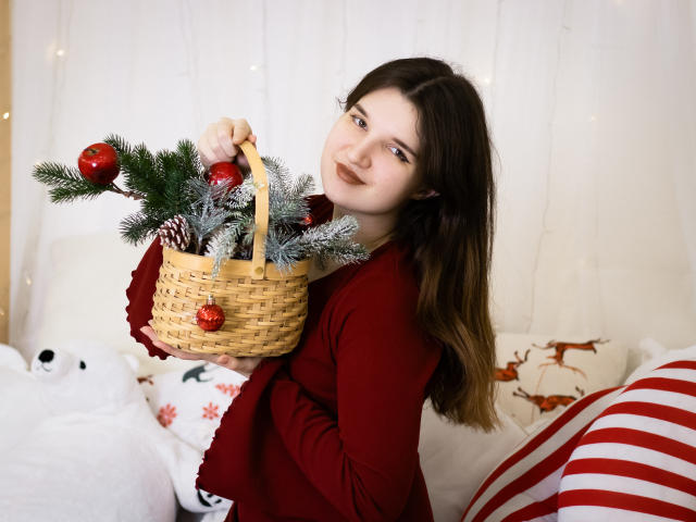 BonnieFlower with long brown hair, wearing a red long-sleeve shirt, holding a woven basket with pine, red ornaments, and a small red ball. She's smiling slightly.