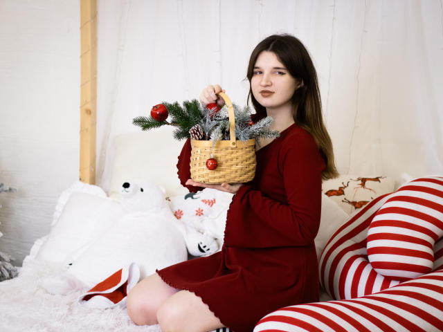 BonnieFlower with medium tits, wearing a red dress, holding a wicker basket with pine and red ornaments, sitting on a striped red and white couch, white pillows.