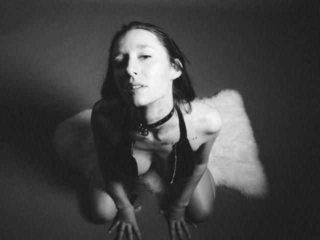 Black-and-white photo of LyllaX69, kneeling, topless, with small tits, wearing a choker, hands on floor, fluffy white rug behind, looking up with parted lips, short dark hair.