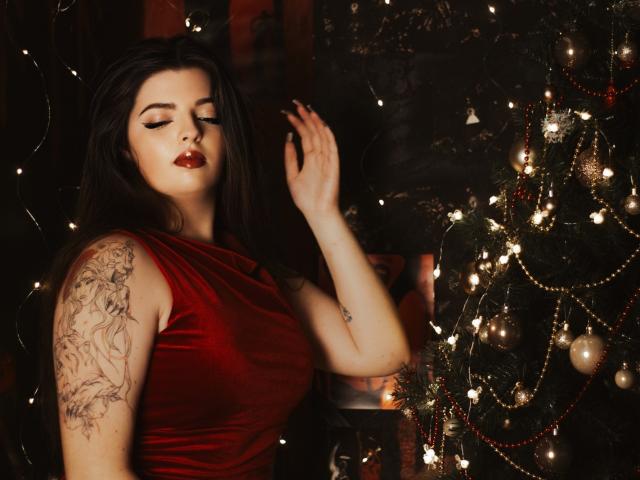 PamDares, avec ses cheveux foncés, sa robe en velours rouge, ses tatouages sur le bras droit, les yeux fermés, la main levée, se tient à côté d'un sapin de Noël décoré de guirlandes et d'ornements. Ses seins sont visibles..