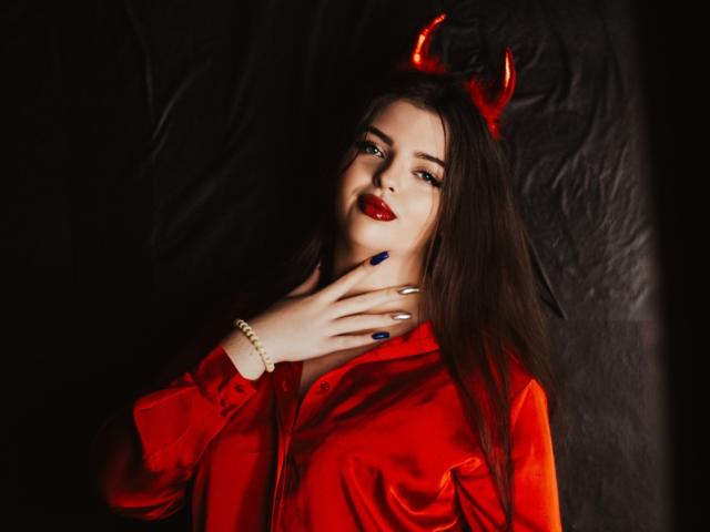 PamDares with red devil horns, red satin shirt, blue nails, white bracelet, and red lipstick, touching her neck, dark background. Her tits are visible, she's looking seductive.