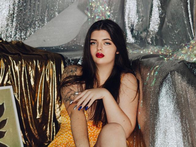 PamDares with long dark hair, red lipstick, and tattoos, sitting in shiny silver and gold fabric, wearing an orange dress, blue nails, looking at camera with confident expression.