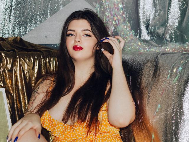 PamDares with long dark hair, red lipstick, wearing an orange polka dot dress, touching her hair, blue nails, sitting on shiny gold and silver-covered surface.