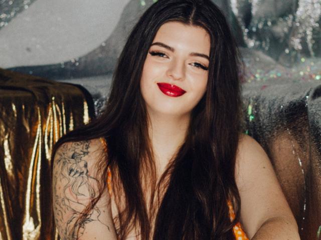 PamDares with long dark hair, red lipstick, and a tattoo on her right arm, wearing an orange dress, sitting in a gold and shiny silver backdrop.