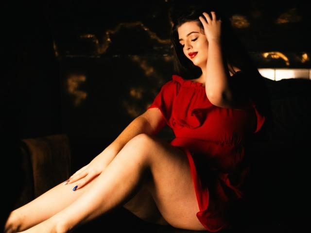 PamDares with red dress, sitting, legs crossed, hand in hair, dark background, red lipstick, blue nails, soft light, curvy, pale skin, showing her ass, relaxed, sexy.