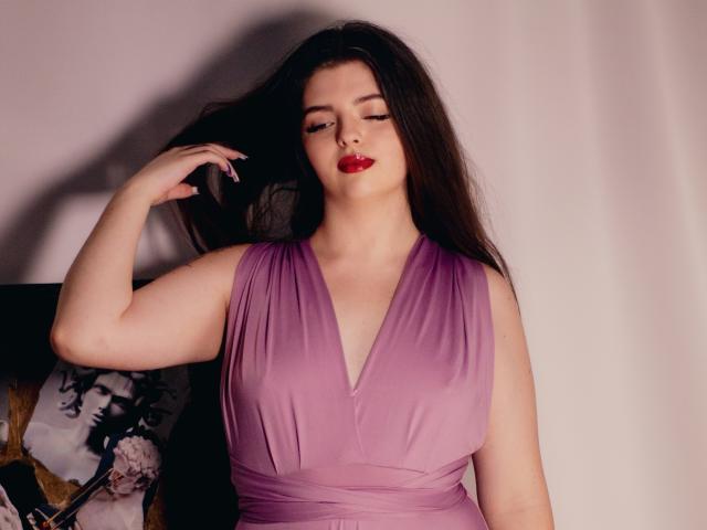PamDares a la peau pâle, de longs cheveux noirs et du rouge à lèvres. Elle porte une robe violette avec un décolleté en V plongeant, et tient ses cheveux d'une main. Sa poitrine est visible..