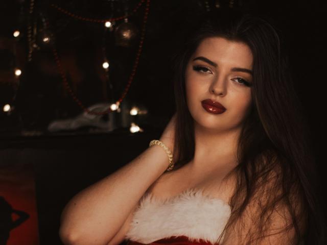 PamDares with long brown hair, red lipstick, and a fur-trimmed red top, holding her hair, wearing a gold bracelet, looking sexy and confident.