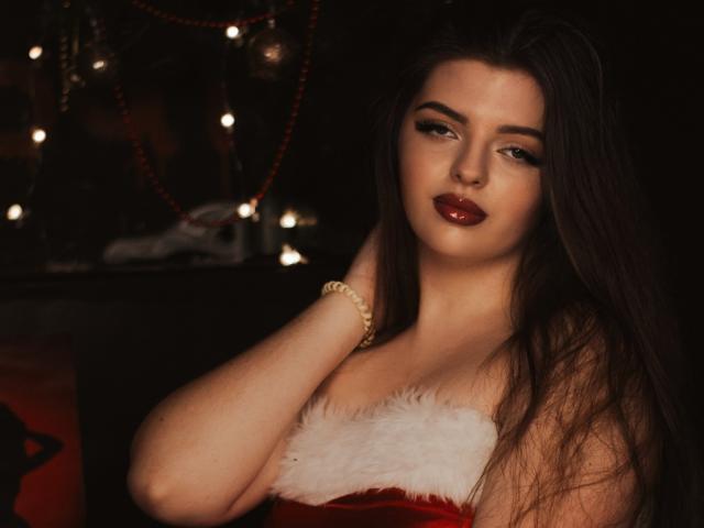 PamDares with long dark hair, red lipstick, and a red velvet dress with white fur, touching her neck, wearing a gold bracelet, looking seductively at the camera.