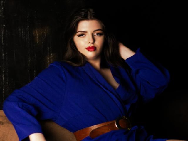 PamDares with long dark hair, red lipstick, wearing a blue dress with brown belt, hand in hair, looking at camera, dark background. Boobs partially visible.