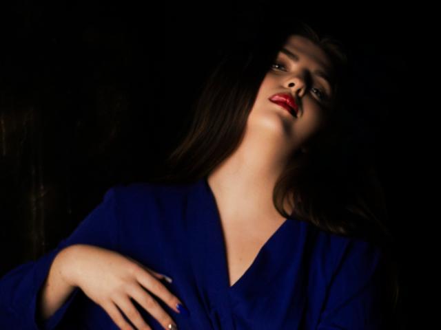 PamDares with red lips, dark hair, and blue robe, looking up, hand on chest, in low light, dark background, showing off her tits subtly.
