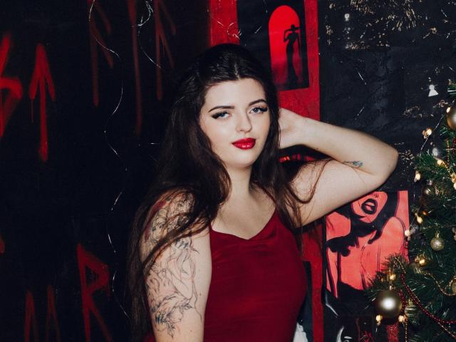 PamDares with long brown hair, red lipstick, and tattoos in a red dress, hand in hair, standing by a Christmas tree, looking at the camera.