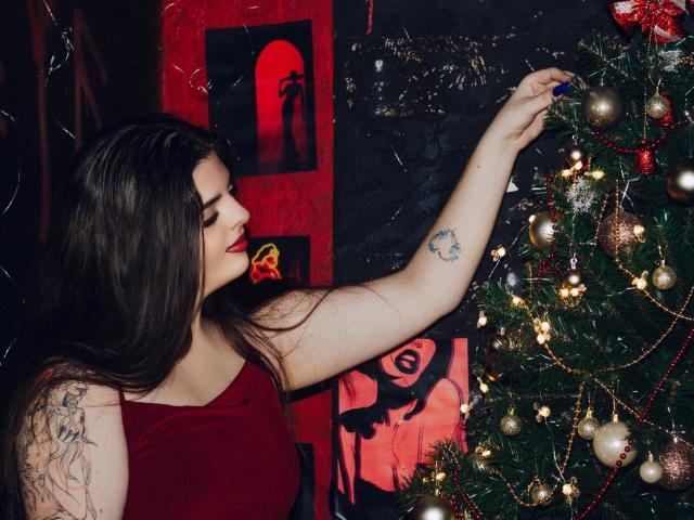 PamDares with long brown hair, red dress, and tattoos, hanging a blue ornament on a decorated Christmas tree, showing a tattoo on her arm.