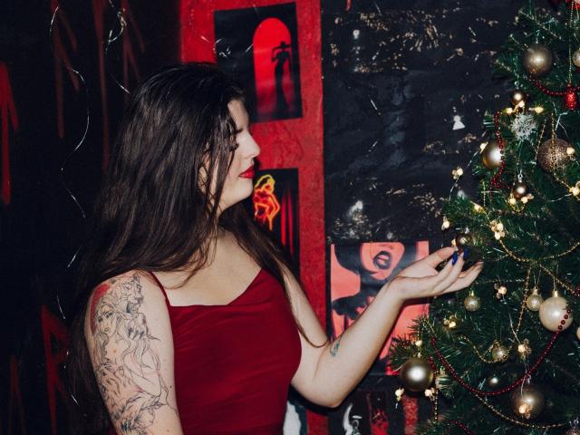 PamDares mit langen braunen Haaren, einem roten Kleid und einem tätowierten Arm, die Weihnachtsschmuck berührt, mit rotem Lippenstift und weißen Nägeln, in einem schwach beleuchteten, rot gefärbten Raum..
