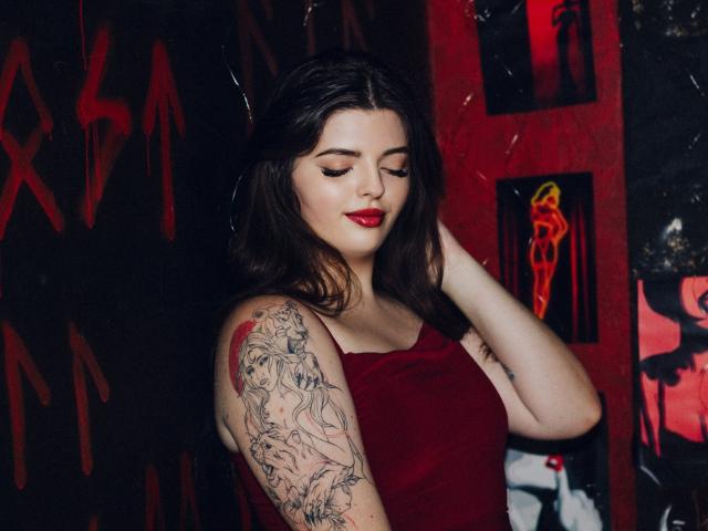 Pam, com batom vermelho, cabelo castanho escuro e vestido vermelho, exibindo o braço tatuado com desenhos intrincados em preto e vermelho, está em pé com os olhos fechados, a mão no cabelo..