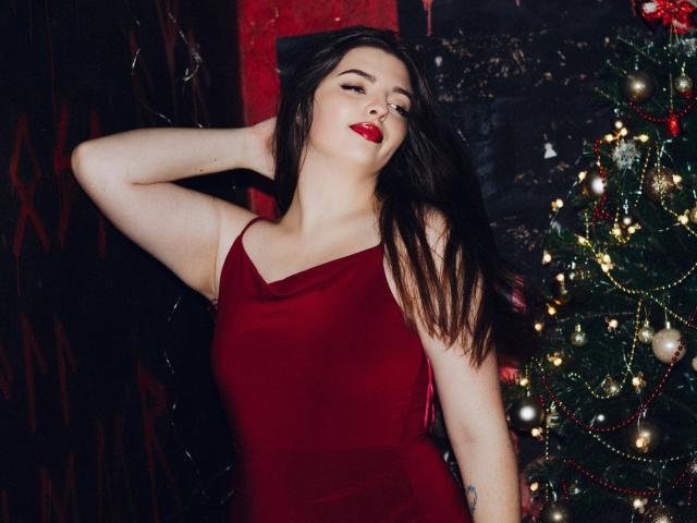 PamDares with long brown hair, red lipstick, and red dress, poses with arm behind head, standing next to a decorated Christmas tree. Her tits are partially visible.