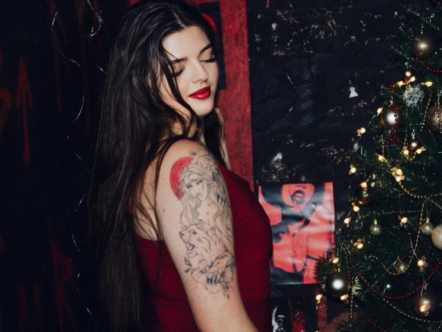 Pam, con cabello largo y oscuro, vestido rojo, lápiz labial rojo, tatuaje en el brazo derecho, de pie junto a un árbol de Navidad con luces, mirando hacia abajo con una ligera sonrisa. Se ven los senos..