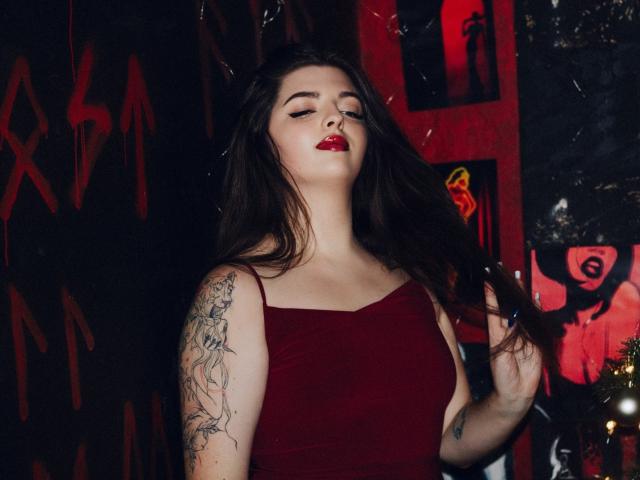 Pam, con lunghi capelli scuri, rossetto rosso e un tatuaggio sul braccio destro, indossa un abito rosso, ha gli occhi chiusi, una mano tra i capelli e sta in piedi di fronte a una parete rosso scuro..