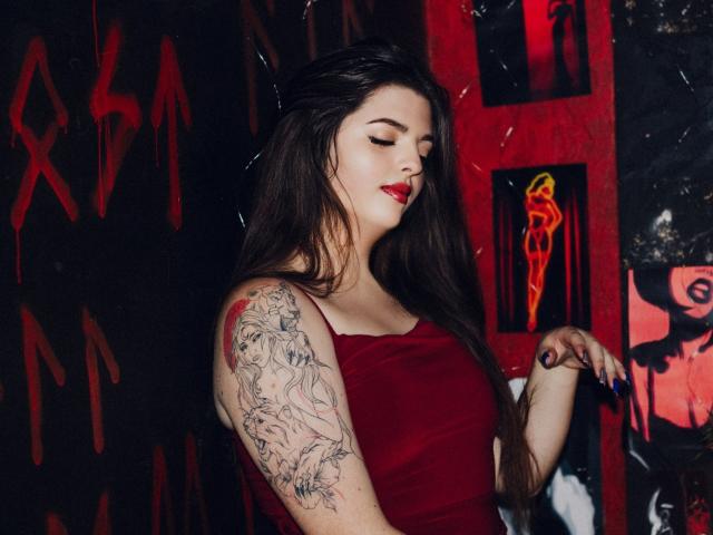 PamDares with long dark hair, red lipstick, and a red dress, showing a tattooed arm, eyes closed, and hand up. Red and black graffiti in the background.